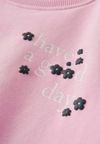 Różowa bluza z teksturowanymi czarnymi aplikacjami w kształcie kwiatów i białym nadrukiem z napisem "miłego dnia". Miękka tkanina z prążkowanym dekoltem.