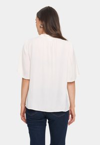Krämfärgad blus med korta ärmar, samlad halsringning och ett subtilt strukturerat mönster, i kombination med mörkblå jeans.