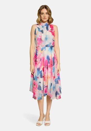 Frau steht und trägt ein ärmelloses, hochgeschlossenes, knielanges Kleid mit Blumenmuster in Pink-, Blau- und Weißtönen sowie weiße offene Absatzschuhe.