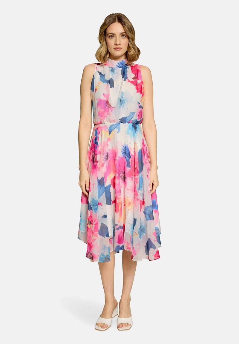 Femme debout portant une robe sans manches, à col montant, longueur genou, à motifs floraux dans des tons rose, bleu et blanc, avec des talons blancs ouverts à l'avant.