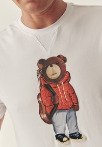 Fehér, rövid ujjú póló, hímzett mintával: egy piros kapucnis pulcsis, kék farmernadrágos medve, aki gitárt tart, és fekete cipőt visel fehér talppal.