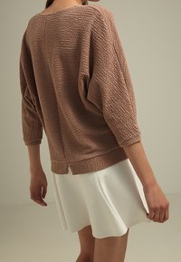 Top marrón texturizado con mangas tres cuartos, ligeramente holgado, combinado con una falda blanca acampanada. Dobladillo visible en la parte trasera.