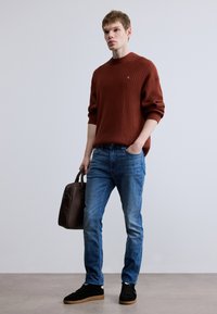 Barna bordázott kötött pulóver, kék farmernadrág, fekete vászoncipő gumitalppal, és barna bőr kézitáska. Egyszerű kialakítás, hétköznapi megjelenés.