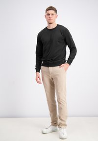 Uomo in piedi con pantaloni beige, maglietta nera a maniche lunghe e sneakers bianche su uno sfondo chiaro e semplice, con una mano in tasca.