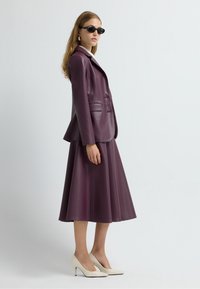 Blazer en cuir bordeaux avec une coupe ajustée et un col en revers, associé à une jupe évasée assortie. Le modèle porte des lunettes de soleil noires et des talons crème.