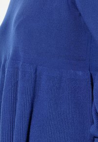 Royalblå stickad topp med texturerad ribbstickning, lös passform och veckade detaljer vid midjan. Tyget verkar mjukt och elastiskt.