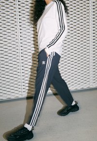White long-sleeve shirt with black stripes on sleeves, paired with dark gray pants featuring three white side stripes and an adidas logo. Black sneakers.