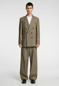 Rjava dvoredna srajca z črnimi gumbi in dvema žepoma, kombinirana z širokimi hlačami in črnimi čevlji. Preprosta bela majica spodaj.