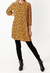 Gul klänning med svart blommönster, rund halsringning och trekvartsärmar, kombinerad med svarta slim fit-byxor och ankelstövlar.