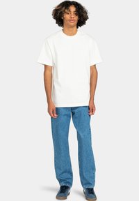 Camiseta de algodón blanca de manga corta, combinada con vaqueros de denim azul holgados. El modelo está de pie sobre un fondo neutro.