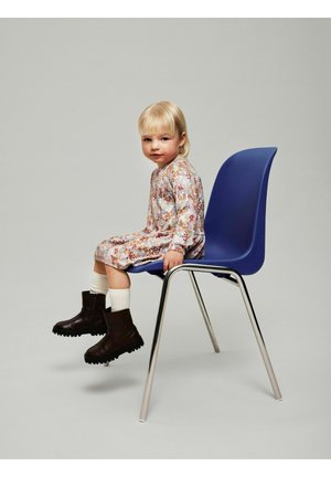 Jeune enfant aux cheveux blonds portant une robe à fleurs, assise sur une chaise en plastique bleu aux pieds en métal, avec des chaussettes blanches et des bottes marron foncé, fond neutre.