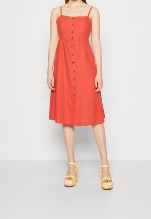 Femme portant une robe midi corail sans manches avec des boutons sur le devant et des sandales compensées jaunes à motif floral.