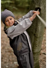 Impermeable y pantalones en gris con acentos claros, con cierre de cremallera, puños elásticos y un bolsillo. Niño tirando de una cuerda sujeta a un árbol.
