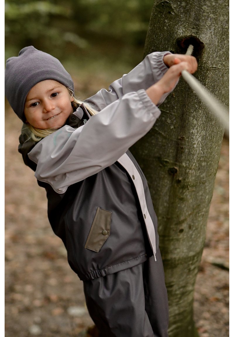 Impermeable y pantalones en gris con acentos claros, con cierre de cremallera, puños elásticos y un bolsillo. Niño tirando de una cuerda sujeta a un árbol.