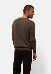 Brauner gestrickter Pullover mit langen Ärmeln, geripptem Saum und Rundhalsausschnitt. Die Textur wirkt weich und hat eine taillierte Silhouette. Getragen mit dunklen Hosen.