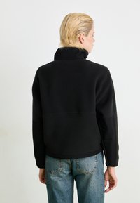 Black fleece jacket with a high collar, featuring a textured, cozy fabric. Paired with blue denim jeans, showing a relaxed fit.