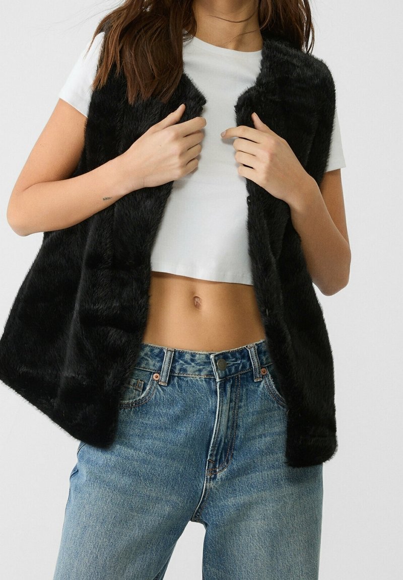 Femme portant une veste en fausse fourrure noire sur un t-shirt blanc court et un jean en denim bleu, mains ajustant l'avant de la veste.