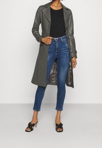 Abrigo de trench de cuero gris con collar muesca, cierre de botones y bolsillos laterales, combinado con jeans ajustados desgastados azules y mule de tacón negro.