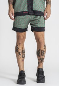 Mann trägt grün-rot gemusterte Shorts und passendes Hemd, dazu schwarze Sneakers und tätowierte Beine sichtbar.
