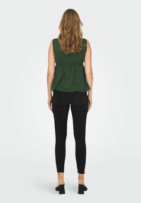 Haut sans manches vert avec un design peplum, associé à un jean slim noir et des sandales à talons carrés. Des motifs et textures simples sont visibles.