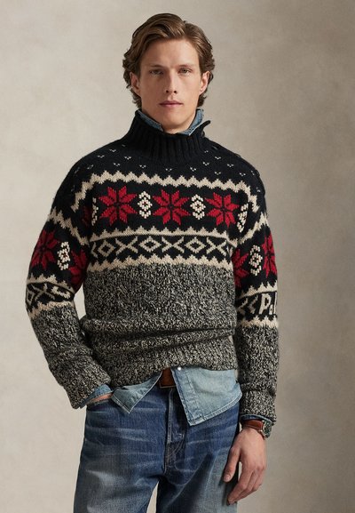 Maglione a maglia in blu scuro con motivi geometrici rossi e crema. Presenta un collo alto e un corpo strutturato. Abbinato a una camicia chambray blu.