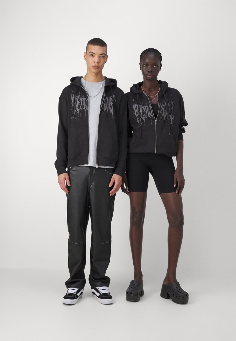 Felpe nere con grafiche, t-shirt grigia, pantaloni in pelle e pantaloni a bicicletta neri, abbinati a sneaker e zoccoli. Sfondo semplice.