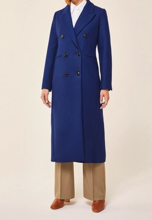 Długi granatowy płaszcz dwurzędowy noszony na białej koszuli, beżowe spodnie o szerokich nogawkach oraz brązowe buty na obcasie na osobie stojącej.