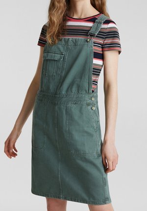 Vihreä denimin haalarihame säädettävillä olkahihnoilla, etutaskulla ja sivunapeilla. Istuva leikkaus ja polvipituinen malli. Raidallinen paita alla.