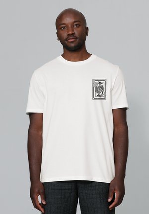 Homme à la peau foncée portant un t-shirt blanc avec un graphisme de carte à jouer joker noir sur le côté gauche de la poitrine, debout devant un fond gris uni.