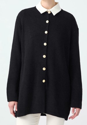 Schwarze, gerippte Strickjacke mit weißem spitzem Kragen und sieben goldenen Knöpfen, getragen über weißen Hosen, gezeigt vom Hals bis zur Hüfte.