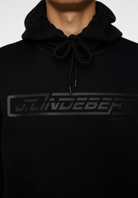 Mustat hupparit, joissa on nyöri, mattamustalla logolla rinnassa ja teksturoidulla kankaalla. Huokuu minimalismia.