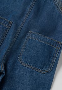 Denimkankaan syvä sininen väri, jossa näkyvät ompeleet; siinä on neliömäinen tasku, jossa on terävä läppä ja sileä pinta.
