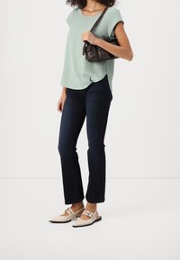 Top court vert clair à manches courtes, jeans bleu foncé et chaussures beiges à bout pointu avec des lanières. Petit sac à main noir avec fermeture rabat.