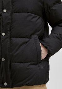 Svart pufferjakke med quiltet design, trykknappknapper, sidelommer og merketag på sømmen; glatt tekstur, isolerende materiale.