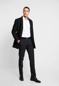 Zwarte blazer over een wit overhemd met zwarte knopen, gecombineerd met donkere broek en schoenen. Soepele textuur en aangesloten ontwerp.