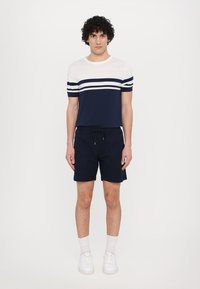 Chemise à rayures bleu marine et blanche à manches courtes avec col rond, associée à un short bleu marine à cordon de serrage. Le modèle porte des baskets blanches et des chaussettes de cheville.