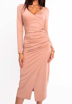 Frau trägt ein langärmliges, gerafftes, rosafarbenes Midikleid mit einem Kreuzanhänger vor weißem Hintergrund.