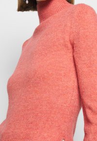 Maglione rosa a collo alto lavorato a maglia con una texture morbida e spacchi laterali; presenta un colletto a costine e una vestibilità leggermente rilassata.