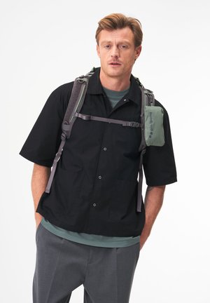 Mann mit schwarzer Kurzarmjacke, grauer Hose, grünem Rucksack mit Brustgurt und angebrachtem grünen Beutel, stehend vor einem einfarbigen Hintergrund.