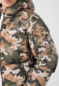 Giacca imbottita mimetica con cappuccio. Presenta una finitura lucida, tasca con cerniera sulla manica e una toppa rotonda con il logo. I colori includono verde e marrone.