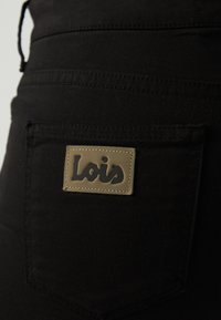 Černé látkové kalhoty s zadní kapsou, na které je hnědý štítek s vyraženým nápisem "Lois" tmavým písmem. Viditelné detaily šití.