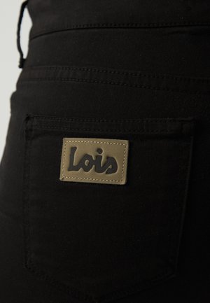 Černé látkové kalhoty s zadní kapsou, na které je hnědý štítek s vyraženým nápisem "Lois" tmavým písmem. Viditelné detaily šití.
