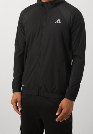 Homme portant une veste noire à capuche avec fermeture éclair complète, un logo blanc à trois bandes sur la poitrine et un pantalon noir assorti.