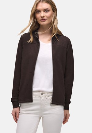 Młoda kobieta w czarnej, teksturowanej kurtce zapinanej na zamek, na białym t-shircie i kremowych spodniach, stojąca na jednolitym tle.