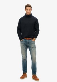 Suéter de punto de cuello alto en azul oscuro combinado con jeans de un azul claro. Un cinturón de cuero marrón y botas de cordones marrones completan el look.