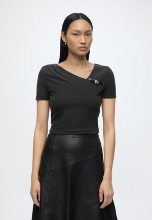 Femme aux cheveux noirs et raides portant un haut noir à manches courtes avec un décolleté asymétrique et une jupe en cuir noire sur un fond uni.