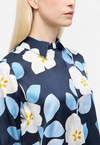 Mørkeblå bluse med store hvide og lyseblå blomster mønstre, gule detaljer og en struktureret krave. Glat stof med en subtil glans.