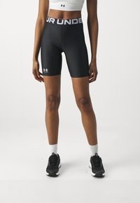 Czarne spodenki sportowe z wysokim stanem z napisem "UNDER ARMOUR" w białym kolorze, wykonane z elastycznego materiału. Noszone z czarnymi sneakersami i białymi skarpetkami.