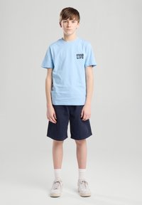 Camiseta de algodón azul claro con "BUENA VIDA" impreso en texto oscuro, combinada con pantalones cortos azul marino. El modelo lleva zapatillas blancas y calcetines blancos.