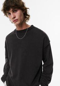 Zwarte oversized sweatshirt met verlaagde schouders, zachte textuur, ribgebreide kraag en minimalistisch ontwerp. Zichtbare zilveren ketting.